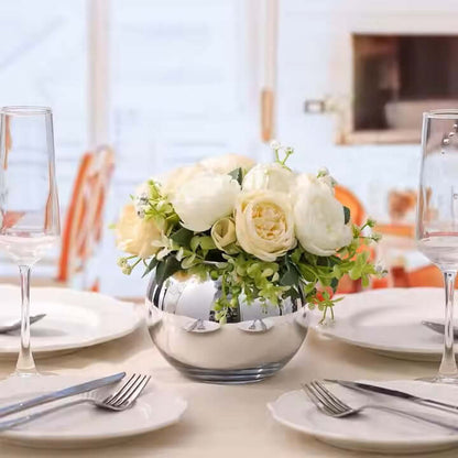 Round fish bowl flowers vase with white roses and greenery on a dining table setting.